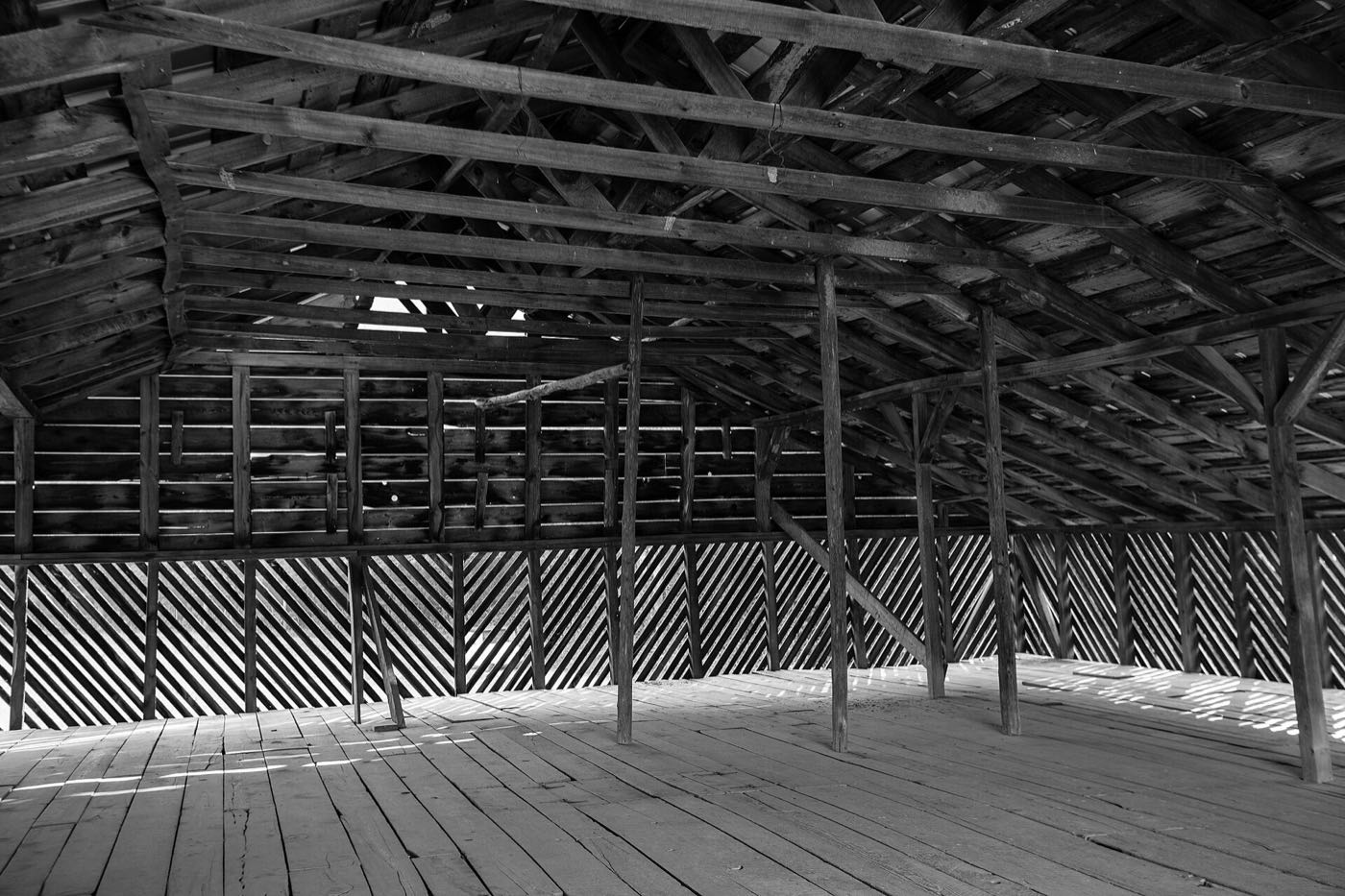 Black-and-white view inside an old timber barn: a plank floor, rows of posts and a heavy roof structure, with daylight through slatted sides