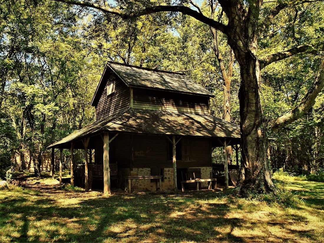 A two-story weathered wooden tobacco pack house with a wide shingled porch roof, shaded by large trees at Duke Homestead in Durham