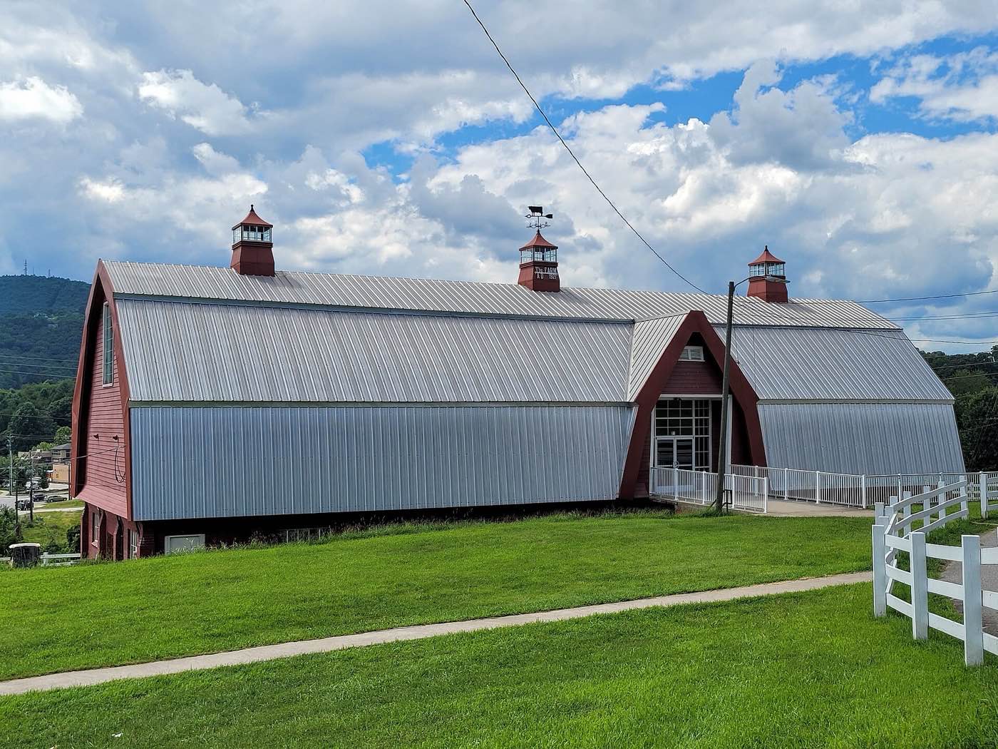 A long dairy barn with a metal gambrel roof, red end walls and three cupolas, behind a white fence on a green lawn in Buncombe County
