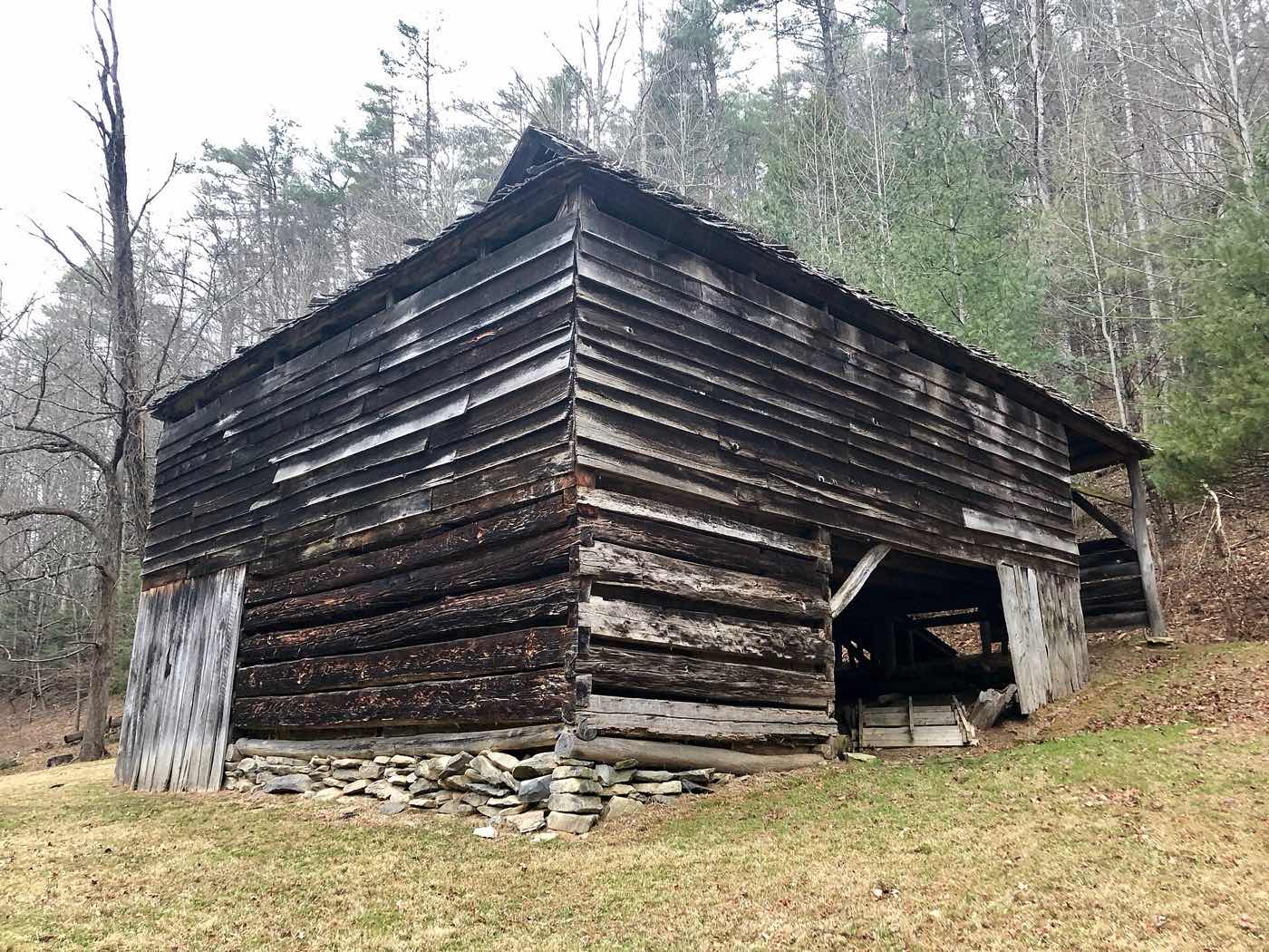 A weathered log and plank barn on a stone footing, with gaps under the eaves and an open side bay, on a wooded slope at Cataloochee